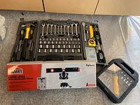 Wide view showing open black plastic tool case with assorted wrenches, sockets, hammer, tape measure, and other tools next to boxed Jobmate laser level and Bossman LiteDriver 2 case.