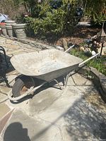 Full view of weathered metal wheelbarrow with single tire and metal handles, placed outdoors on stone paving.