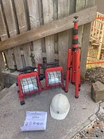 Photo showing the red Globe dual lamp work light with black protective cages, red tripod stand, white hard hat and instruction manual on concrete surface against wooden fence.