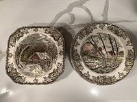 Photo showing two Friendly Village plates, one square with a covered bridge in a snowy village scene, the other round with a stream and trees in a village scene, both have ornate floral borders.