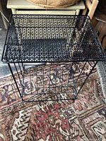 Front view of black metal side table with lattice patterned top and magazine rack below on a patterned rug.