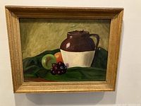 Full view of framed vintage still life painting with crock and fruit on a green cloth and yellowish background.