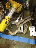 View of yellow handheld work light with black cord and switch next to metal locking pliers clamps and C-clamps on a table.