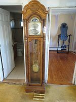 Front view of tall wooden Howard Miller grandfather clock showing ornate carved top, glass door with visible brass pendulum and weights, and clock face with Arabic numerals.