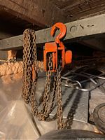 Orange metal chain hoist with multiple chains and large lifting hook suspended from a beam in a basement area