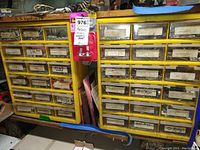 Yellow hardware storage drawer units with visible labels indicating contents such as roll pins, pins, washers, pop rivets, o-rings, and screws. Packaged pull pins attached on front.