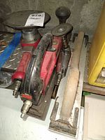Red sander with black handle and round sanding pad next to a wooden handle sanding block, all dusty and used.