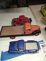 Top view of three toy truck models placed on a white surface including a red vintage tow truck with detached front, an orange Ajax flatbed truck, and a blue pickup truck.