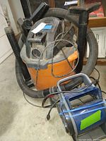 Photo showing Rigid wet/dry vacuum with hose attachments and blue Lasko inflatable fan placed on floor, highlighting condition and cords.