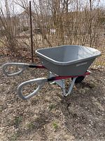 Side angle showing gray plastic basin, red frame, and gray coated handles of the wheelbarrow outdoors on dirt ground.