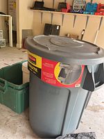 Close-up of Rubbermaid Brute 44 gallon commercial trash can with lid and one green plastic bin behind it in garage.