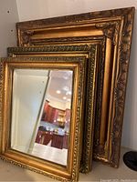 Three gold framed mirrors stacked and leaning against the wall showing ornate detailing on the frames and reflective surfaces.