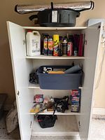 Wide view of white wooden cabinet shelves holding various automotive fluids, aerosol sprays, oil pan, blue bin with tools, and car jack with carrying bag at the bottom shelf.