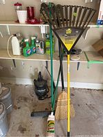 Ames Pro leaf rake, brooms, mop, garden hose, cleaning products and black vacuum tool on floor in garage with shelving behind them