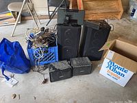Wide view showing two large Bose speakers, two smaller Magnavox speakers, and a blue milk crate of cables alongside boxes in a garage setting.