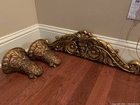Pair of gold tone carved corbels and flat carved arched plaque resting on wooden floor against wall