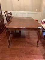 Front view of rectangular wooden dining table with polished wood finish, Queen Anne legs, and silverware drawer handle visible.
