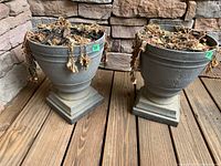 Three plastic urn style flower pots with dried plants inside sitting on a wooden porch floor against a stone wall.