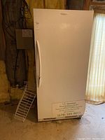 Front view of the upright freezer showing white textured finish, curved handle, and a sign attached to the bottom front panel.
