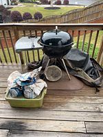 Weber kettle grill on tripod with two wheels, side shelf visible, bags of charcoal and mesquite chips, black grill cover on porch deck.
