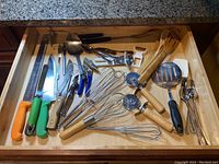 Photo showing drawer filled with various kitchen utensils including different knives, whisks, ladle, can opener, and other kitchen tools.