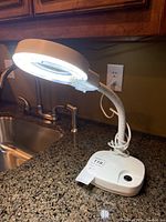 Desk lamp switched on, showing bright LED ring light surrounding magnifying glass, placed on granite countertop near sink.