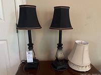 View of two matching table lamps with black trimmed shades and two extra off-white shades on wooden surface by door.