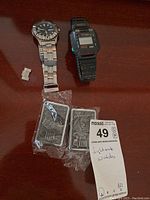 Two American Legend collectible lighters in plastic wrap alongside two wristwatches on a wood surface, one analog silver band watch and one black digital watch.