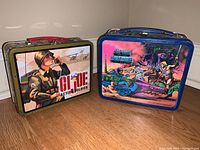 Two vintage metal lunchboxes side by side showing front artwork: GI Joe soldier image and He-Man fantasy battle scene.