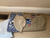Lot items including folded beige bucket hat, khaki IZOD pants with Flex Stretch tag, and blue checkered IZOD button-up shirt still in plastic packaging.