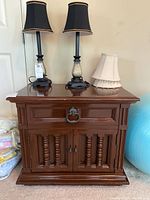 Front view of wooden nightstand with two lamps and one fabric lamp shade on top, showing drawer and cabinet door with decorative spindle accents and metal ring pulls.