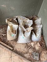 Eight white plastic bags filled with pea pebbles and stones standing against a concrete wall with some debris on the ground around them.