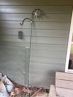 Full view of tall black metal double shepherd's hook leaning against a wooden siding. Shows overall height and structure with suet box attached to one side.