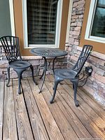 Full set of wrought iron outdoor table and two matching chairs positioned on wooden porch floor against stone wall.