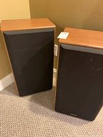 Pair of Technics wood veneer speakers with black grilles standing on carpet, viewed from front and angled side