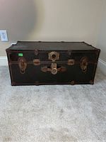 Front angled view of a rectangular metal trunk with rusted metal latches and dark exterior on carpeted floor.