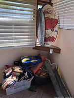 Photo showing boogie boards, baseball mitts, sports shoes, hockey and lacrosse equipment clustered in corner near window blinds.