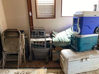 Photo showing three large coolers stacked on top of each other in blue, teal, and white colors, with additional camping gear including a rolling basket and sleeping bags behind.