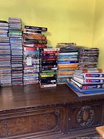 Photo showing large stacks of CDs, DVDs, and books on a dark wooden sideboard against yellow wall. Items are piled in multiple columns including boxed sets and single cases.