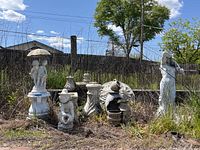 Wide view of several plaster/cement garden statues and bird baths outdoors on grass with fence in background