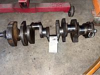Full length view of the Chevy 350 small block engine crankshaft showing the crank journals, counterweights, and gear on one end. The crankshaft is resting on a concrete floor beside a red vehicle lift.