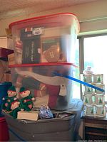 Two large clear plastic bins with red lids containing assorted Christmas decorations. Three elf dolls and other small holiday items outside the bins on a gray container.