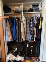 Wide view showing a variety of men's medium and large, and women's small, medium, large clothing. Includes shirts, coats, jeans, gloves, and luggage at bottom. Clothes hung on wooden rod and folded items on shelves above.