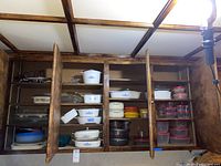 Photo showing CorningWare and Pyrex serving bowls with plastic storage containers inside a wooden cabinet with open doors.