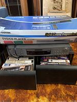 Photo showing Toshiba DVD/CD player SD-K720 in original box along with VHS tapes and black speaker cover.