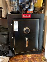 Closed front view of black metal safe with Edelbrock logo plaque, dial combination lock, and handle with auction tag attached.