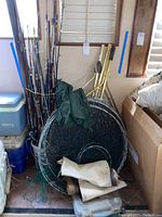 Overall view of fishing rods bundles, fishing nets, waders, and tackle box stacked together against a wall indoors.