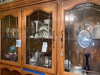 Wide view of oak wood china cabinet with glass doors showing inside shelves containing various glassware, china plates, cups, and figurines.
