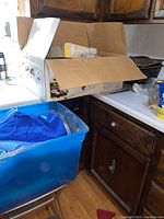 Partially opened cardboard box and blue plastic tote containing vintage roaster pans and other kitchenware on countertop