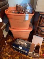 Two stacked orange plastic bins labeled 'Xmas' filled with various holiday decorations including baskets and smaller boxes on the floor next to them.